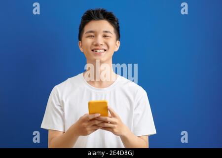 Casually dressed student standing isolated on blue background looking at screen of smart phone, browsing web pages and smiling while chatting with fri Stock Photo