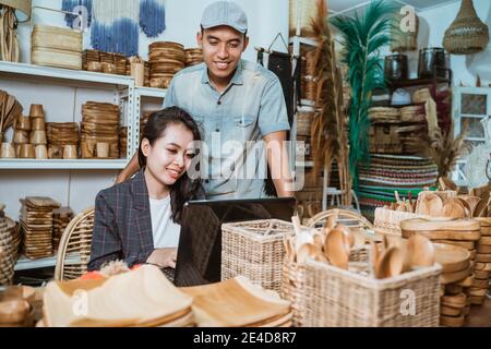 a male businessman standing next to a female secretary who sits while working on a laptop at a handicraft workshop Stock Photo
