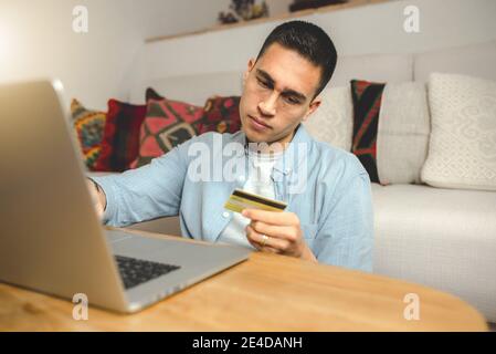 Young Hispanic man holding a credit card for payment using a laptop computer at home Stock Photo