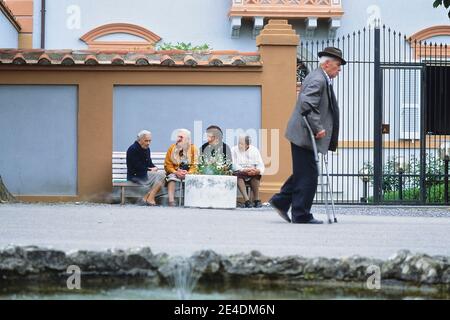 An elderly man with crutches passing four elderly Italian ladies sitting on a public bench having a discussion. Italy Stock Photo