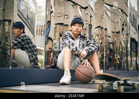 teenage asian kid taking a break sitting on ground with basketball and skateboard Stock Photo