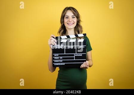 Young beautiful brunette woman pregnant expecting baby over isolated yellow background holding clapperboard very happy having fun Stock Photo