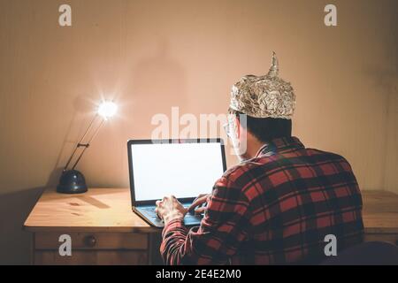 Young man with aluminum cap is sitting in the dark basement in front of a laptop. Conspiracy theory concept Stock Photo