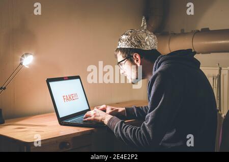 Young man with aluminum cap is sitting in the dark basement in front of a laptop. Conspiracy theory concept Stock Photo