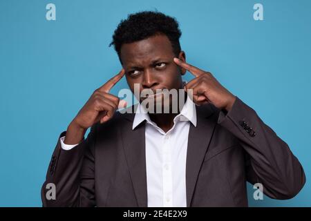 Puzzled african guy in suit tries to recollect important information in mind, keeps fore fingers at temples poses against blue studio background Stock Photo