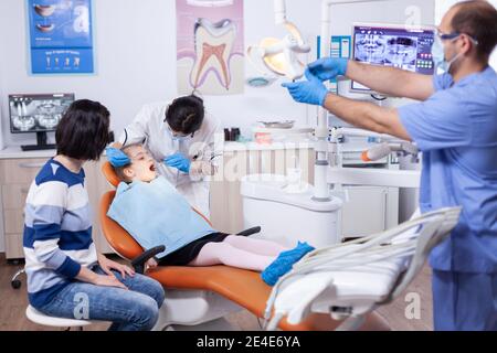 Kid patient in dental office getting teeth treatment sitting on dental chair wearing bib with mouth open. Dentistry specialist during child cavity consultation in stomatology office using modern technology. Stock Photo