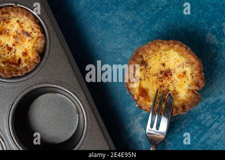 A freshly baked egg custard tart viewed from above with a cake for resting on the tart. A baking tray showing another tart is also visible. On a stone Stock Photo