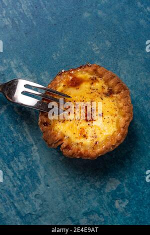 A freshly baked egg custard tart viewed from above with a cake fork resting on the tart. The tart is on a stone blue background Stock Photo