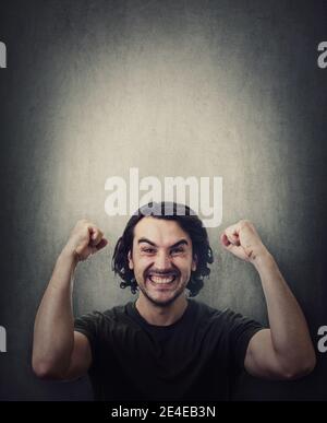 Pleased and positive man, long curly hair style, celebrates success hysterical and passionate. Guy keeps fists tight, raise hands up clenching teeth, Stock Photo