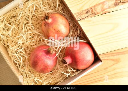 Three organic pomegranates, close-up, in a box with shavings on a wooden table. Stock Photo