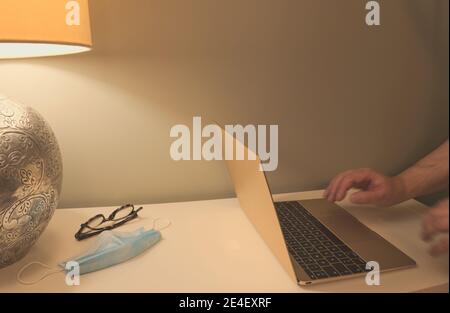 Hands of an unrecognizable man working and typing on laptop with a mask, glasses on the table Stock Photo