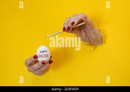Happy Easter abstract theme. Hands painting a white easter egg while breaking through cutouts of a yellow cardboard background Stock Photo
