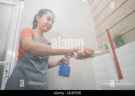 smiling Asian woman wipes using a window wiper and holds the bottle sprayer while cleaning the toilet glass in the bathroom Stock Photo