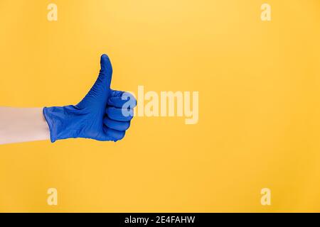 Close up of male in medical protective blue gloves makes thumb up gesture, demonstrates approval or agreement, isolated on yellow wall with copy space Stock Photo