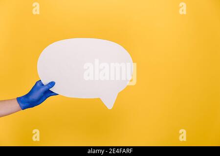 Close up of unrecognizable female in medical protective gloves hold white paper blank speech bubble, isolated on yellow studio background Stock Photo