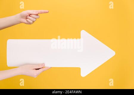 Close up of female hands holding big white paper arrow, finger pointing at copy space for advertisement, isolated on yellow studio background Stock Photo