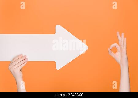 Unrecognizable female holding white paper arrow points to OK gesture, demonstrates approval or agreement, isolated on orange background with copy spac Stock Photo