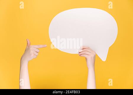 Close up of unrecognizable young female holding and pointing at blank white paper speech bubble, isolated on yellow background with copy space Stock Photo