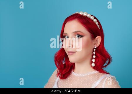 Close-up face of a young woman with red wavy hair in a white dress, pearl headband and long dangling earrings, isolated on a blue background Stock Photo