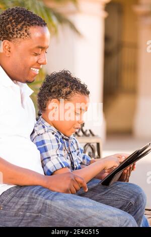 African American Father and Mixed Race Son Using Computer Tablet on Bench in Park. Stock Photo