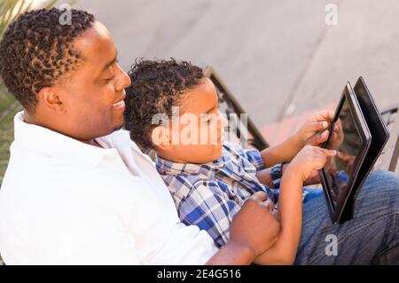 African American Father and Mixed Race Son Using Computer Tablet on Bench in Park. Stock Photo