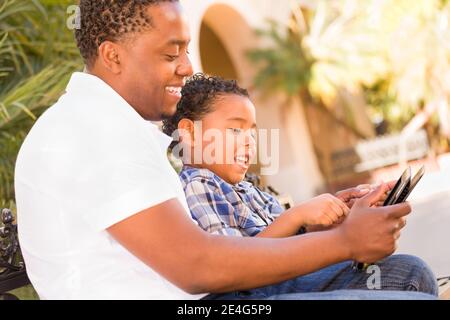 African American Father and Mixed Race Son Using Computer Tablet on Bench in Park. Stock Photo