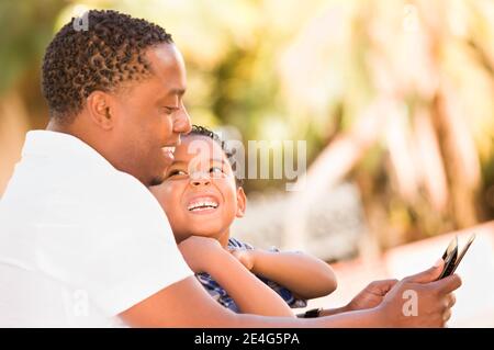 African American Father and Mixed Race Son Using Computer Tablet on Bench in Park. Stock Photo