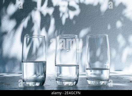 Three glasses of water standing on a white rim against a bright white wall with shadows of leaves on a sunny summer day Stock Photo