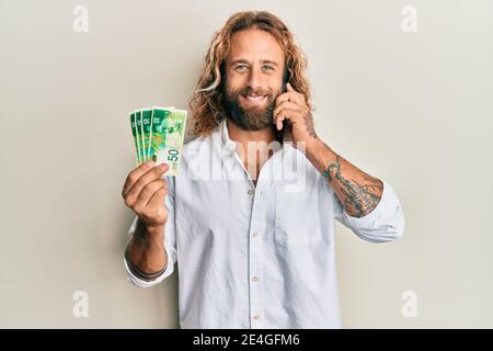 Handsome man with beard and long hair talking on the phone holding 50 shekels smiling with a happy and cool smile on face. showing teeth. Stock Photo