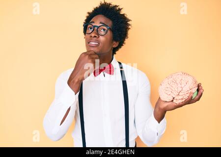 Handsome african american nerd man with afro hair holding brain serious face thinking about question with hand on chin, thoughtful about confusing ide Stock Photo