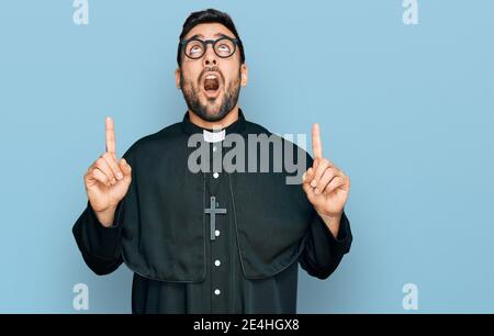Young hispanic man wearing priest uniform amazed and surprised looking up and pointing with fingers and raised arms. Stock Photo