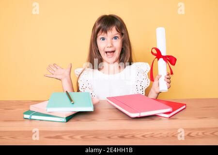 Little caucasian kid girl with long hair holding graduate degree diploma for preschool celebrating achievement with happy smile and winner expression Stock Photo