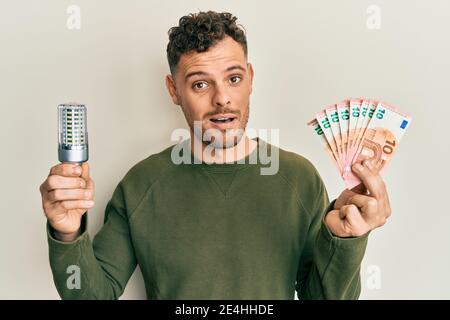 Young hispanic man holding led bulb and euros banknotes in shock face, looking skeptical and sarcastic, surprised with open mouth Stock Photo