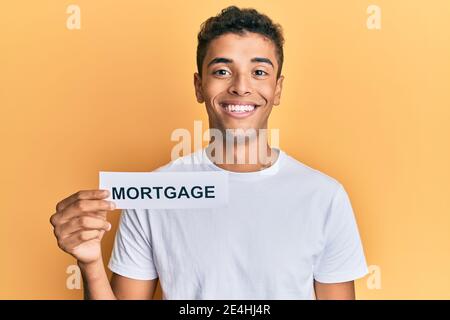 Young handsome african american man holding mortgage word looking positive and happy standing and smiling with a confident smile showing teeth Stock Photo