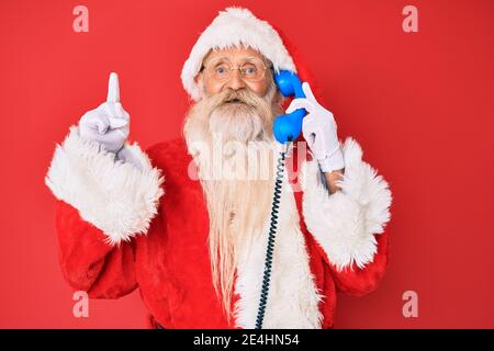 Old senior man wearing santa claus costume using vintage telephone surprised with an idea or question pointing finger with happy face, number one Stock Photo