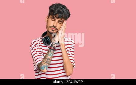 Young hispanic man listening to music using headphones sleeping tired dreaming and posing with hands together while smiling with closed eyes. Stock Photo