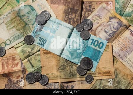 One hundred reais (current brazilian currency) displayed at the top of former currencies of Brazil with coins Stock Photo