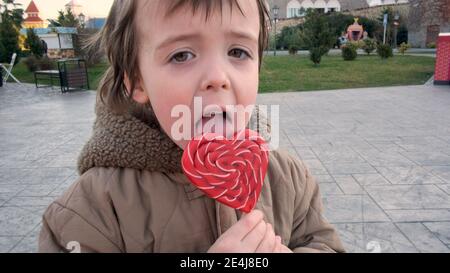 Funny little boy in warm brown jacket licks large heart shaped lollipop spending time in city park on autumn day closeup Stock Photo