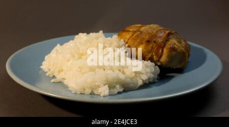 Boiled white rice with chicken cutlet wrapped in bacon on a gray background blue plate. Baked chicken meatball with garnish. Stock Photo