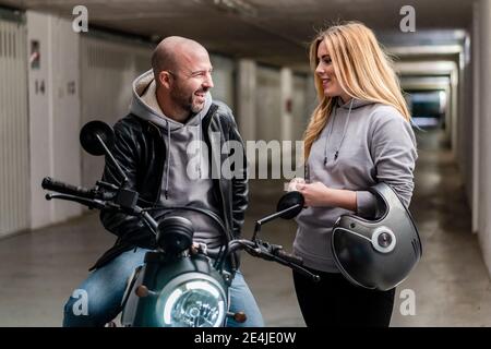 Smiling woman talking with boyfriend sitting on motorcycle in parking lot Stock Photo