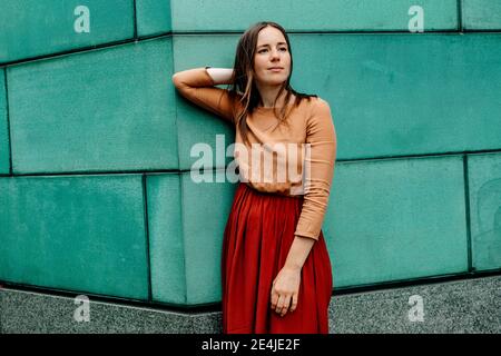 Woman with hands behind head contemplating while looking away against green wall Stock Photo