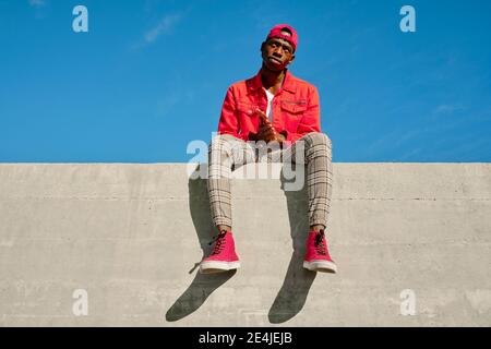 Portrait of young man in red jacket and baseball cap sitting on concrete wall Stock Photo