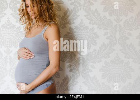 Pregnant woman touching belly while standing against wall at home Stock Photo