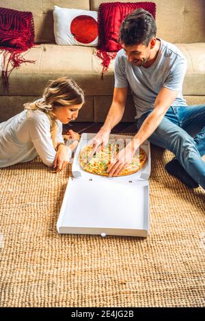 Young couple sitting on floor of living room and cutting pizza in box Stock Photo