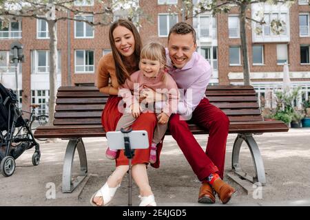 Happy family with one daughter talking selfie from mobile phone on tripod at park bench Stock Photo