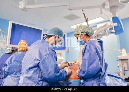 Doctors wearing face mask operating surgery while standing with colleague in background at operation room Stock Photo