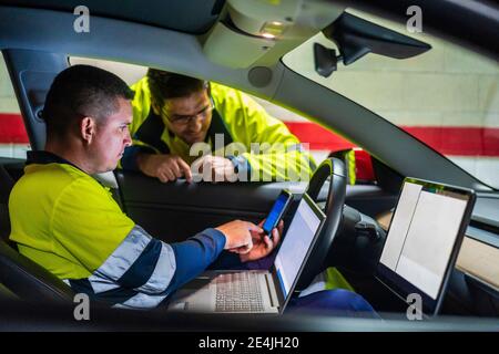 Male programmer showing mobile phone to colleague while sitting in electric car Stock Photo
