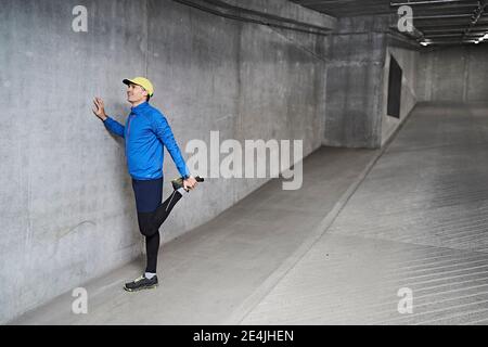 Smiling mature man stretching leg by gray wall in parking garage Stock Photo