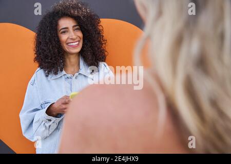 Smiling young woman talking with female friend while standing against wall Stock Photo