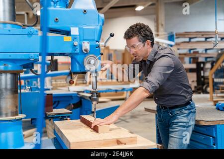 Carpenter wearing protective goggles drilling wooden plank in production hall Stock Photo